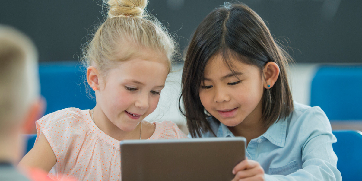 Duas meninas sentadas na sala de aula, uma asiática e uma loira sorriem enquanto olham para um tablet.