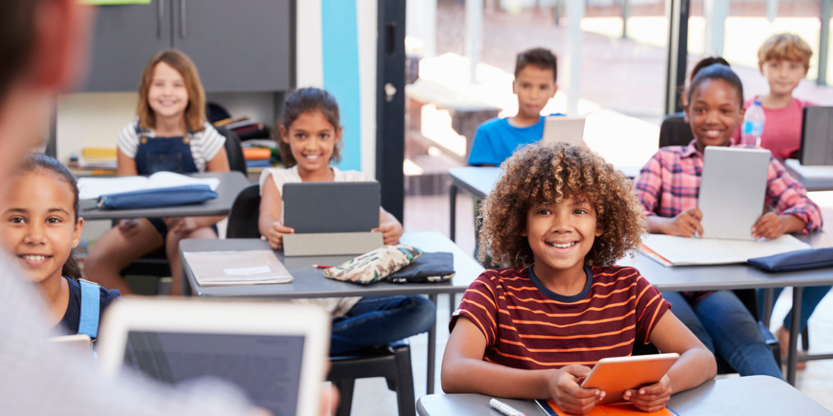 Sala de aula com alunos diversos com tablets na mão acompanhando a explicação da professor.