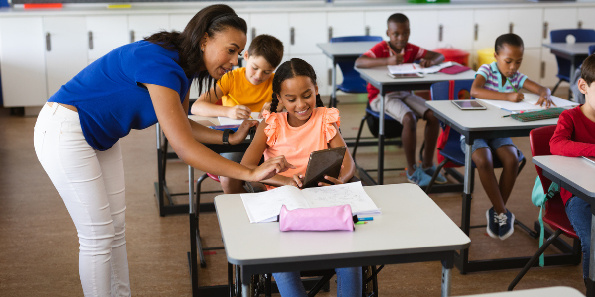 Professora aponta para tablet que esta na mão de aluna, em uma sala de aula com alunos diversos.