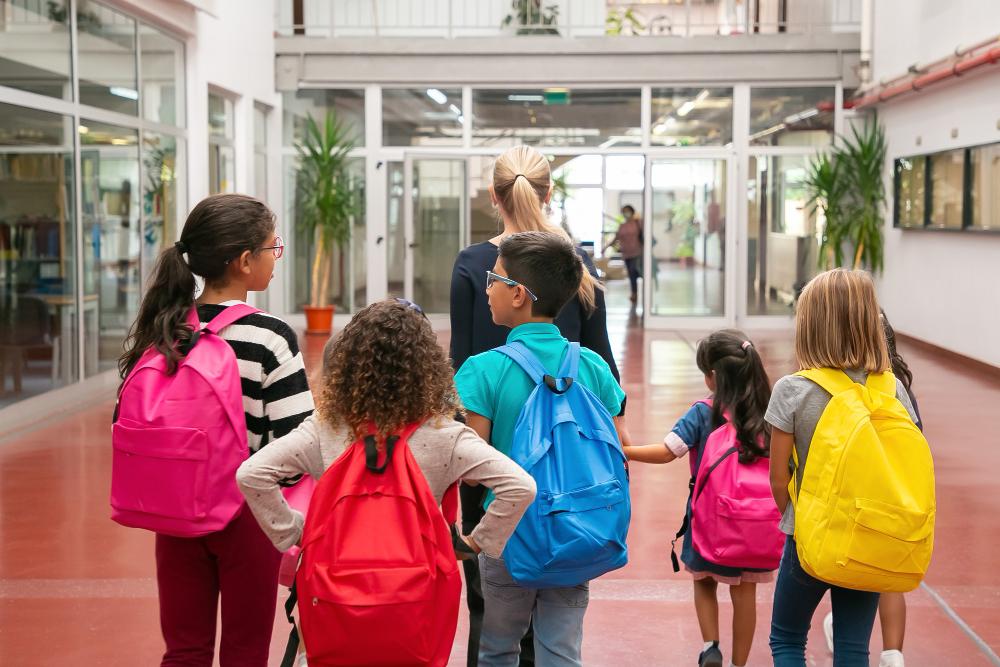 Crianças carregando mochilas enquanto acompanham professora pela escola.