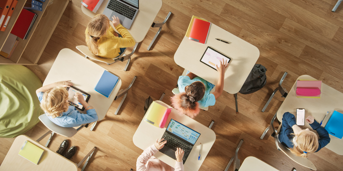 Vista superior da sala de aula no ensino fundamental: crianças sentadas na mesa da escola usando computadores pessoais e tablets digitais para tarefas.