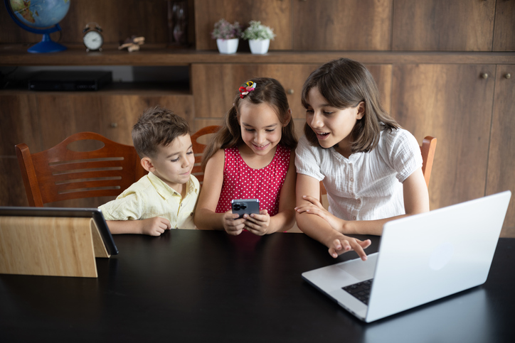 Three children with diverse ages using laptop computer, smartphone and digital tablet at home and having fun