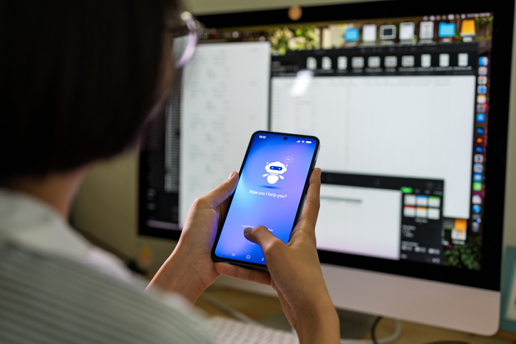 An Asian teenage student interacts with an AI chatbot on a smartphone while studying at a desk with a computer, notes and stationery. The scene highlights modern learning and technology integration.