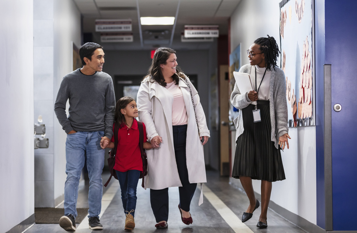 Parents bringing girl to school, with teacher in hallway Two parents bringing their daughter to school, talking with an elementary school teacher or principal in the hallway. Perhaps it's the first day of school, or they are going to have a parent-teacher conference. The multiracial girl is 6 years old. Her father is Asian and her mother is Hispanic.