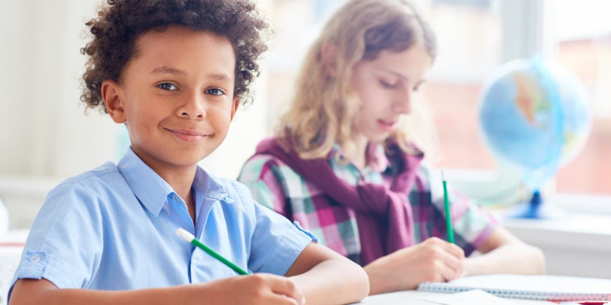 criança sorri com lápis na mão em escola com outra criança ao fundo