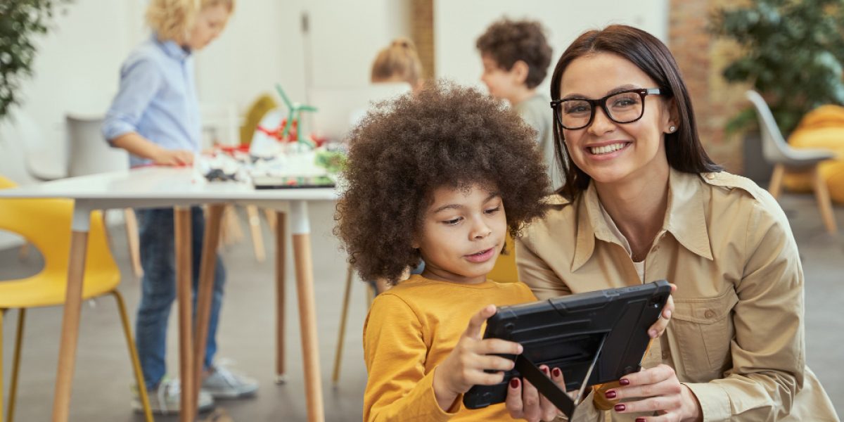 coordenadora pedagógica auxilia um estudante infantil a manusear um tablet.